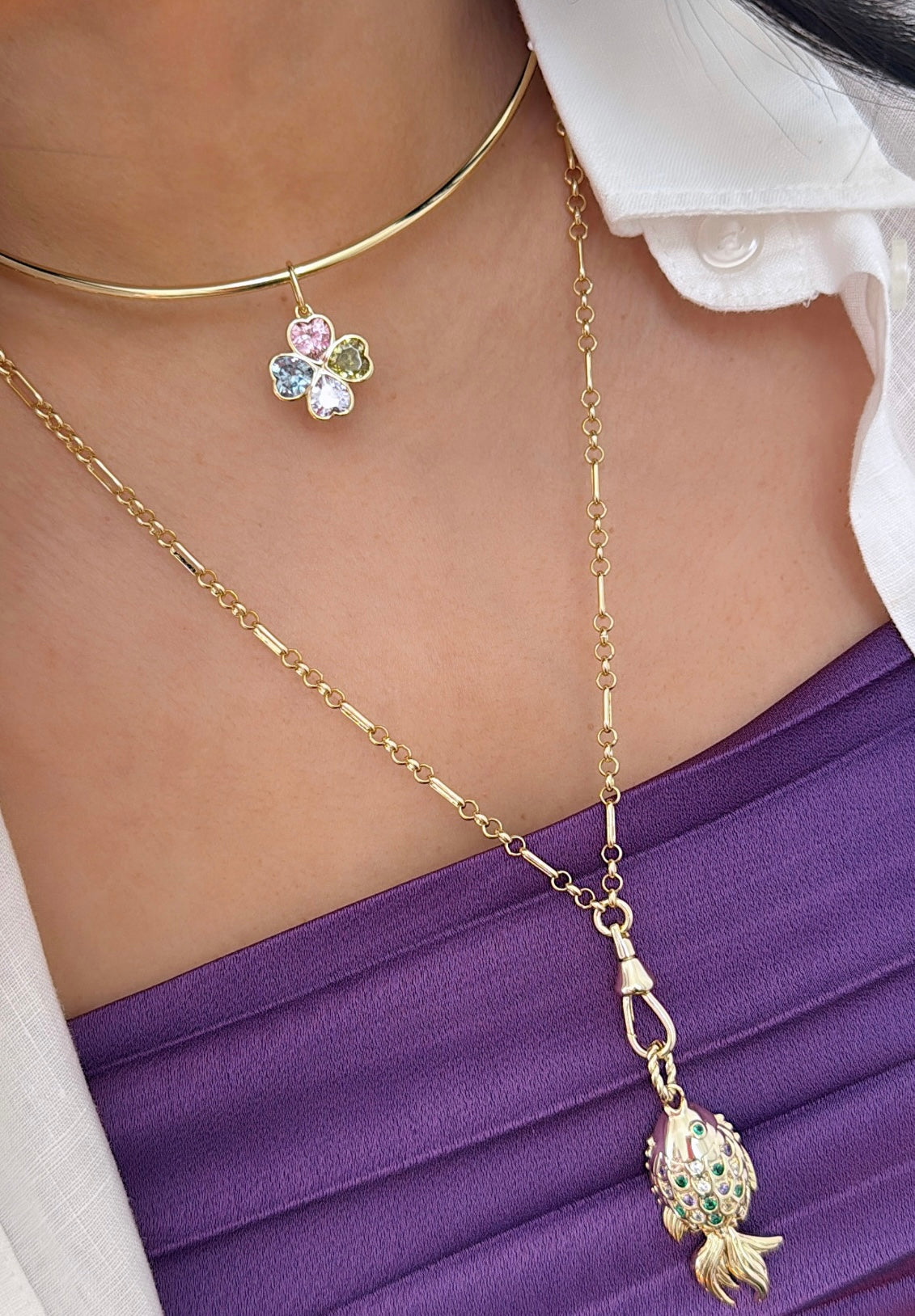 Layered gold necklaces on a woman's neck: a rigid choker with a pastel gemstone four-leaf clover charm and a longer gold chain with a gold fish charm adorned with gems, over a purple top.